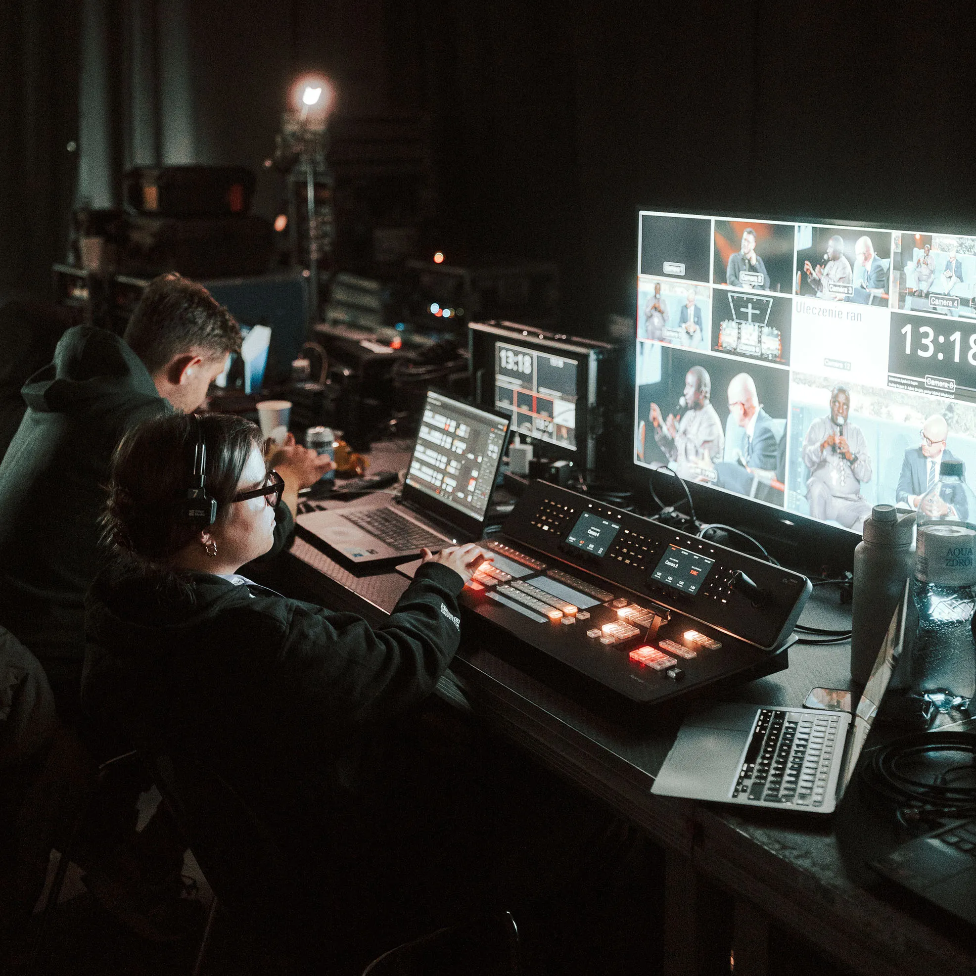 Two people operating video production equipment with monitors displaying a live panel discussion in a dimly lit control room.