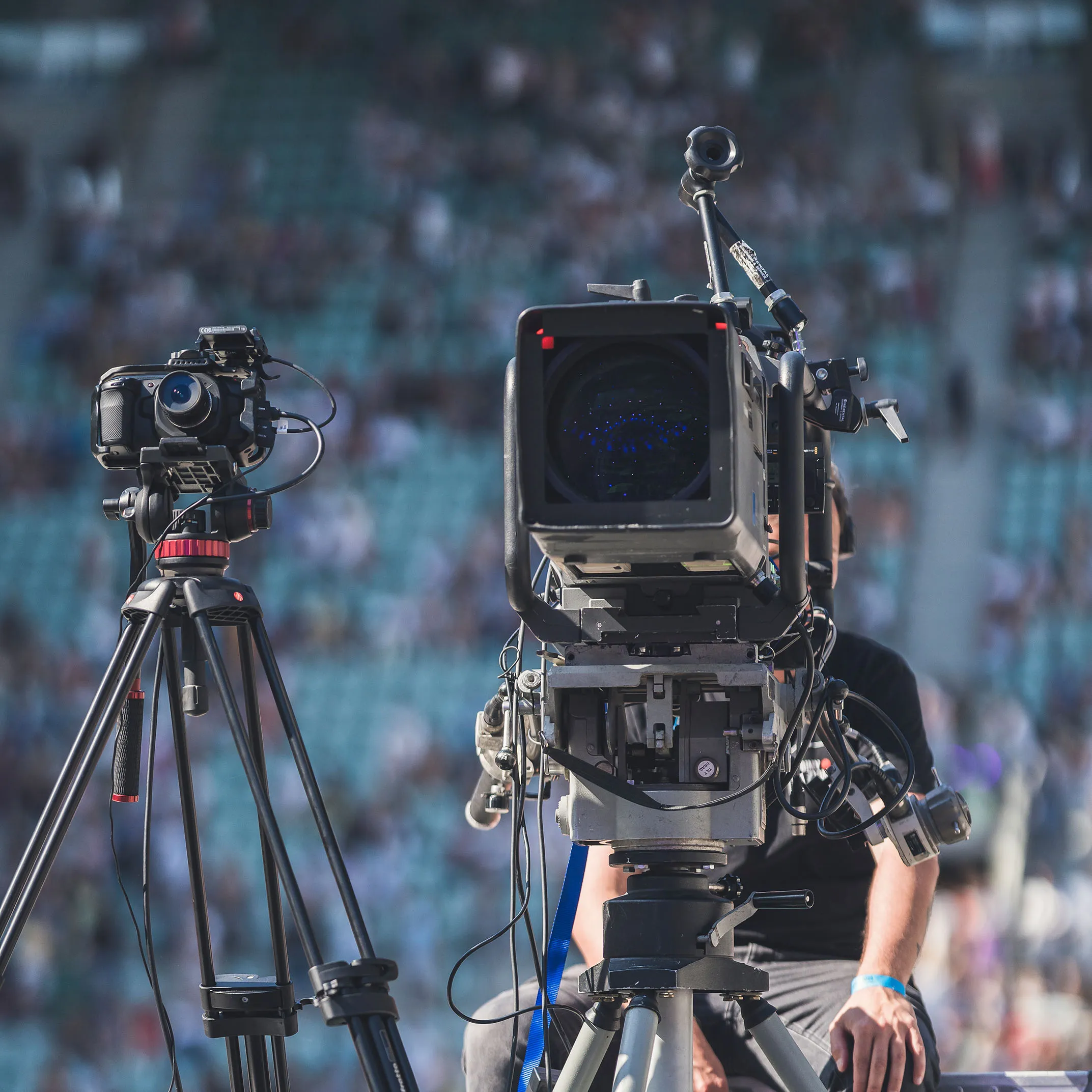 Operator filming with professional video camera on tripod at outdoor event with blurred crowd in background.