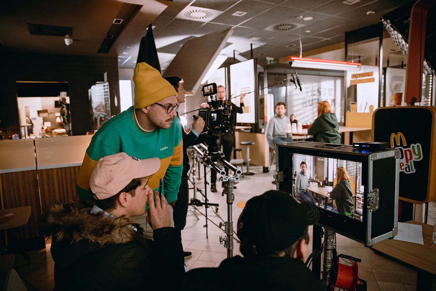 Crew filming a scene with two people sitting at a table inside a fast food restaurant, shown on a monitor.