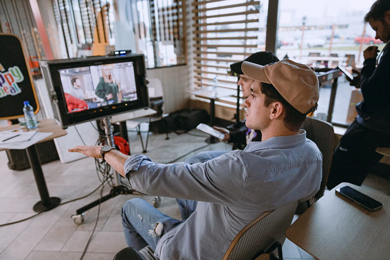 Two men seated indoors watching a monitor showing people in discussion, one man gesturing with his hand.