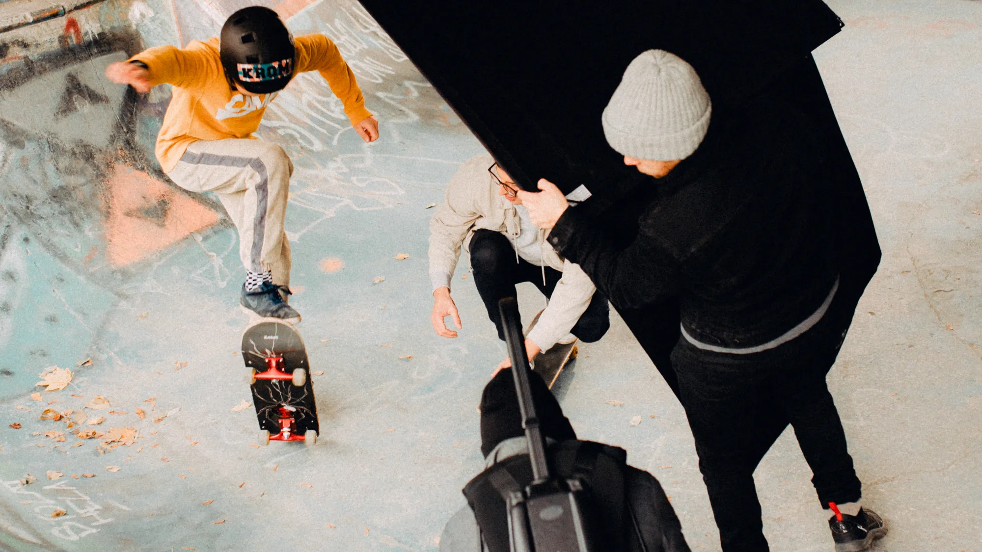 Skateboarder in a yellow sweatshirt performing a trick while three others film or watch in a graffiti-covered skatepark.