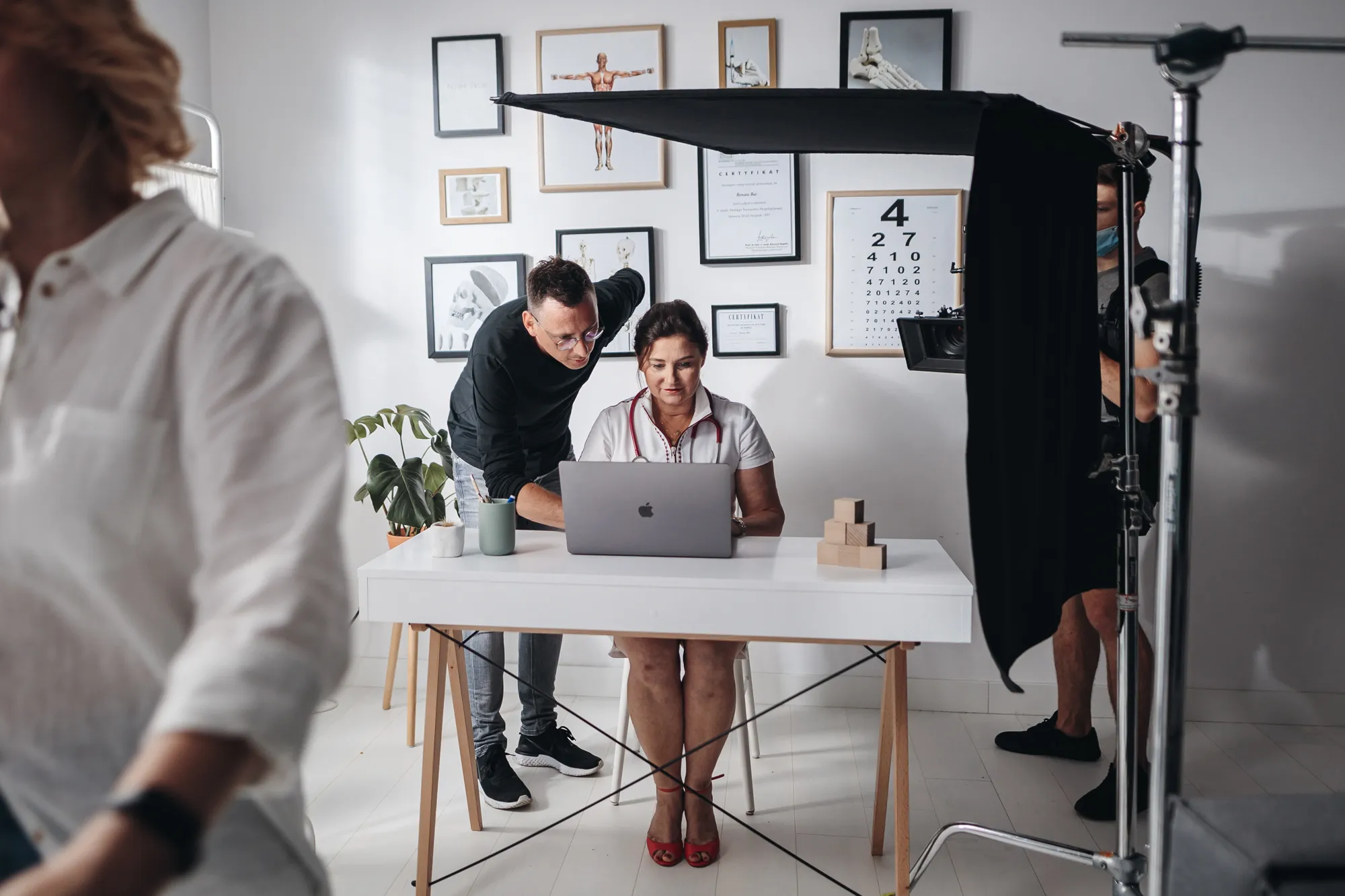 Woman doctor sitting at a desk with a laptop, a man standing next to her pointing at the screen, medical diagrams on the wall, and a cameraman filming behind a black reflector.