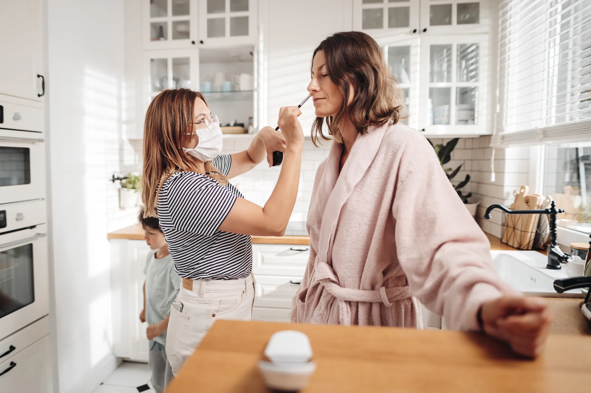 A woman wearing a face mask applies makeup to another woman in a pink robe in a bright kitchen, with a boy standing in the background.