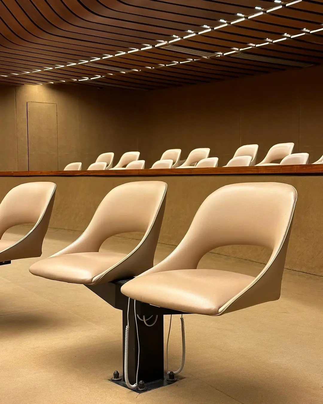 Rows of beige, modern, swivel chairs with a curved design arranged in a room with matching beige walls and carpet and a ceiling with horizontal linear lighting.