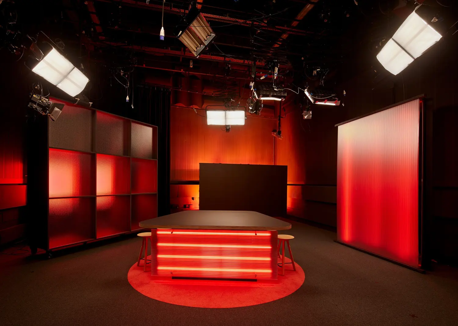 Empty studio set with a table illuminated by red horizontal lights, two wooden stools, red-lit backdrop panels, and overhead studio lights.