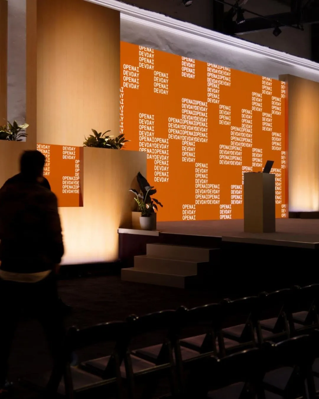 A dimly lit stage setup for a presentation or event with an orange background screen displaying repeated white text 'OPENAI DEVDAY'. There are potted plants on stage and in front, a podium with a laptop, and empty chairs in the foreground. A person is walking at the left side of the image, silhouetted against the light.