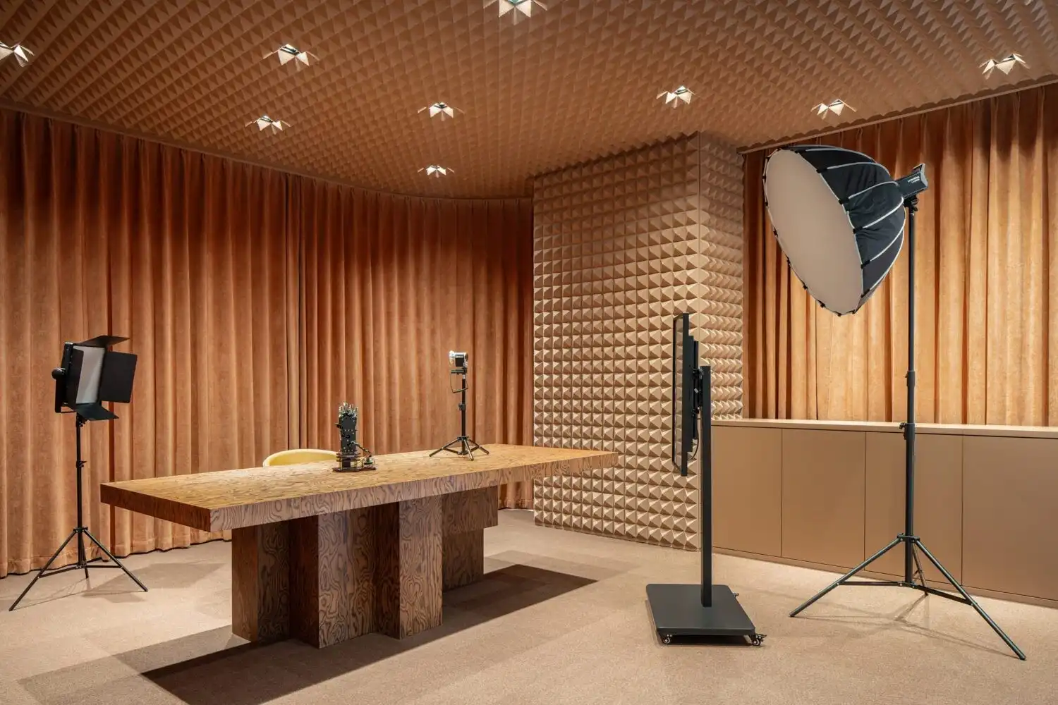 Interior view of a soundproofed room with padded walls and ceiling, featuring a large wooden table with objects on it, a yellow chair, and professional photography lights on stands surrounding the table.
