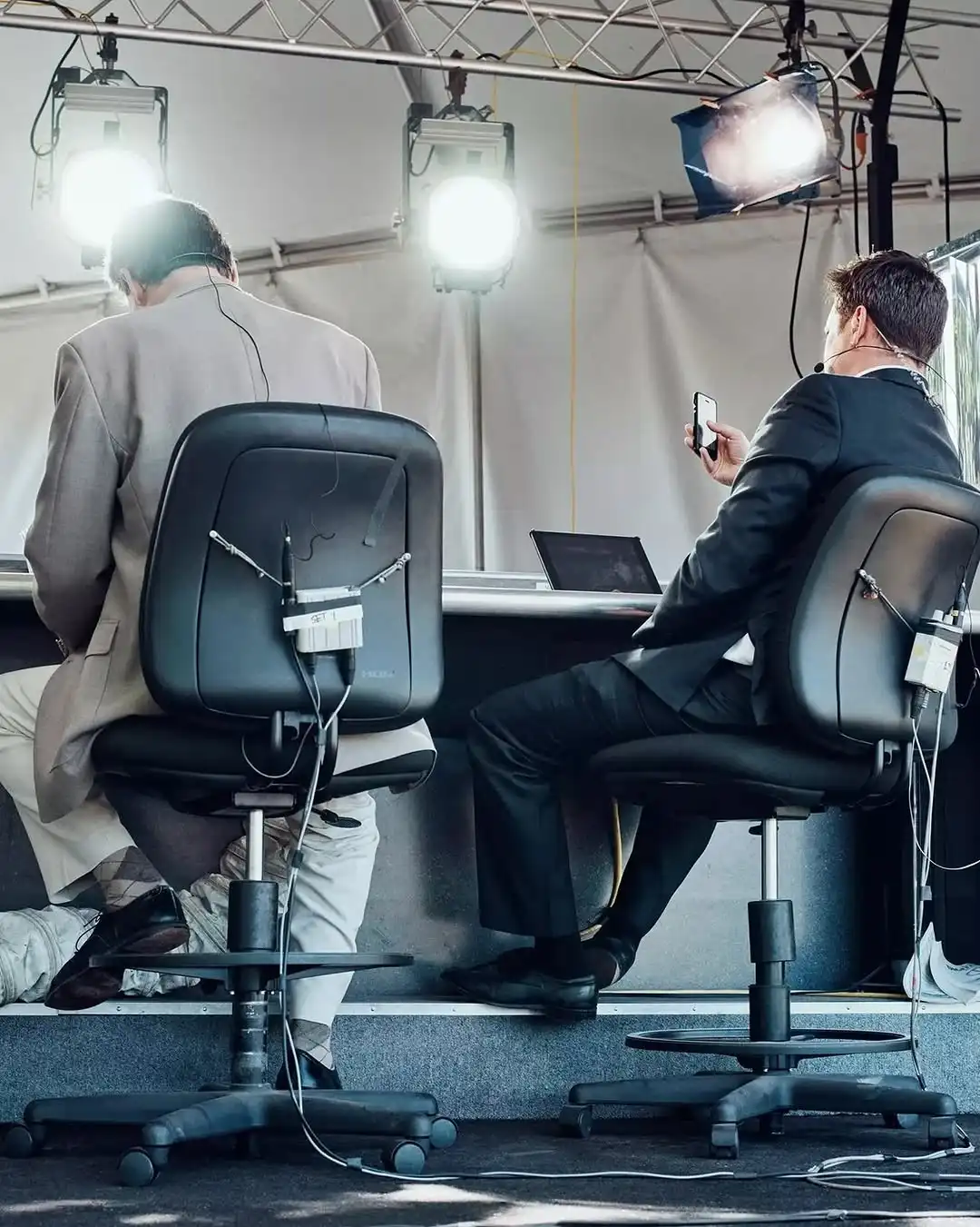 Two men sitting on office chairs with broadcast microphones and wires attached, facing a desk with bright studio lights overhead and one man looking at his phone.