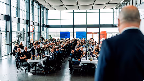 Large group of people seated at tables in a modern, bright hall listening to a bald man in a suit speaking.
