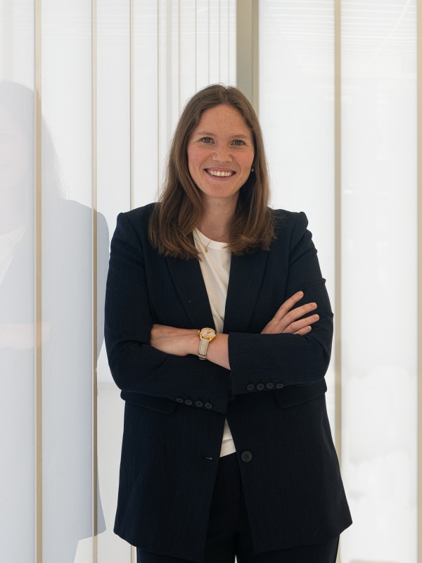 Professional woman in a dark blazer and white shirt smiling confidently with arms crossed, standing by a glass wall in a modern office setting.