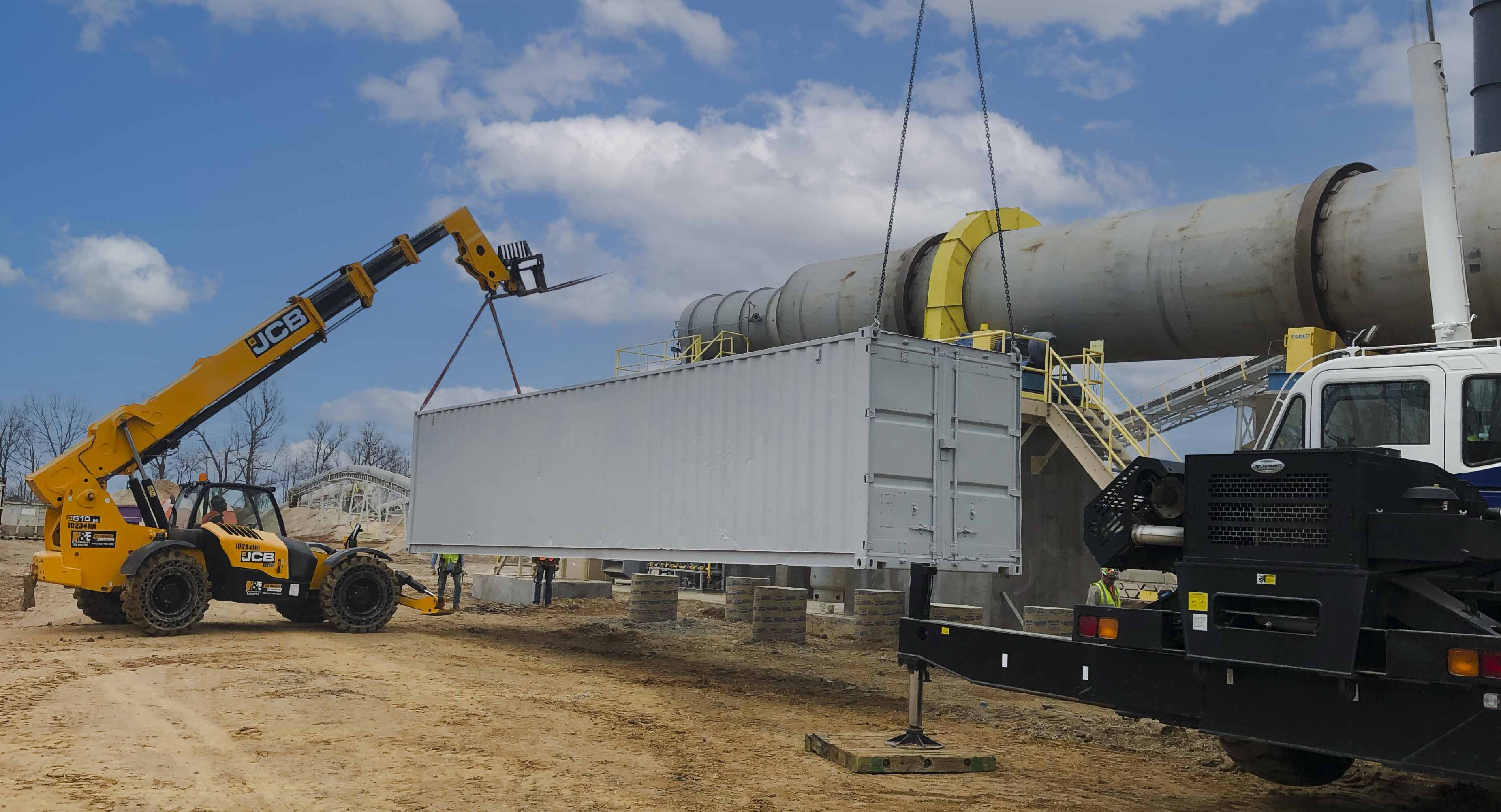Telescopic forklift and crane lifting a shipping container at industrial oilfield site under blue sky