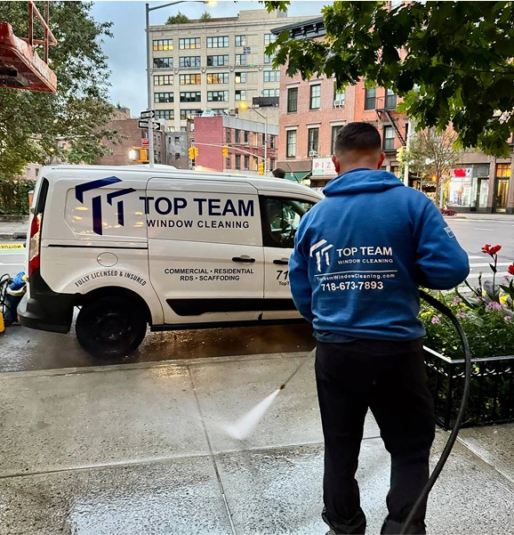Person in a blue hoodie pressure washing a sidewalk next to a white van with Top Team Window Cleaning branding parked on a city street.
