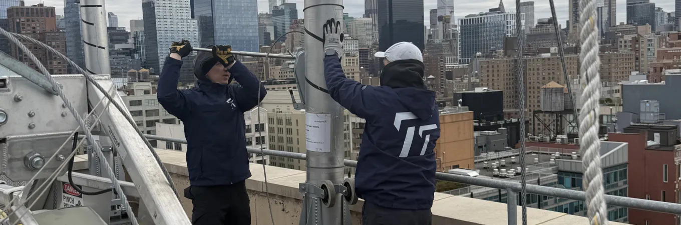 Two workers in jackets and gloves adjusting ropes on a rooftop with city skyline in the background.