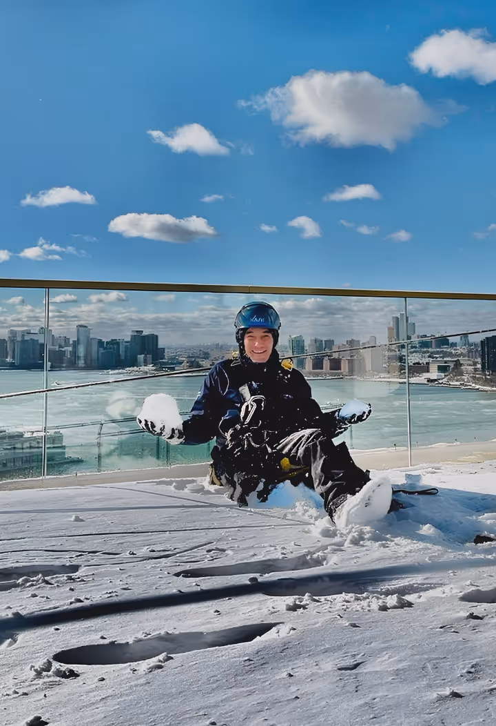 Person in winter gear sitting on snow-covered rooftop with city skyline and blue sky in the background.