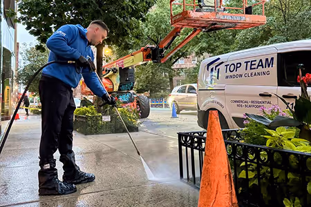 Worker in a blue jacket pressure washing a sidewalk near a Top Team Window Cleaning van and orange safety cone.