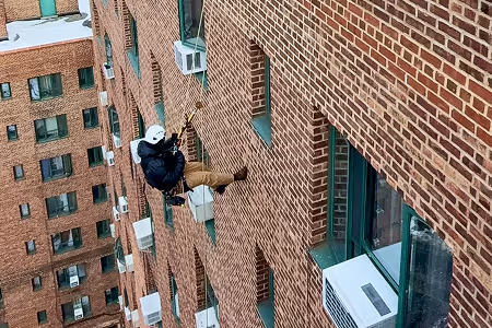 Person wearing safety gear and helmet inspecting or cleaning windows on the exterior of a tall brick building while suspended by ropes.
