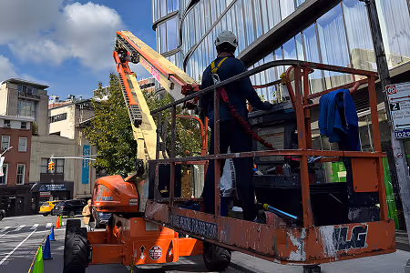 Worker on an orange aerial lift platform performing maintenance on the exterior of a glass building under a partly cloudy sky.