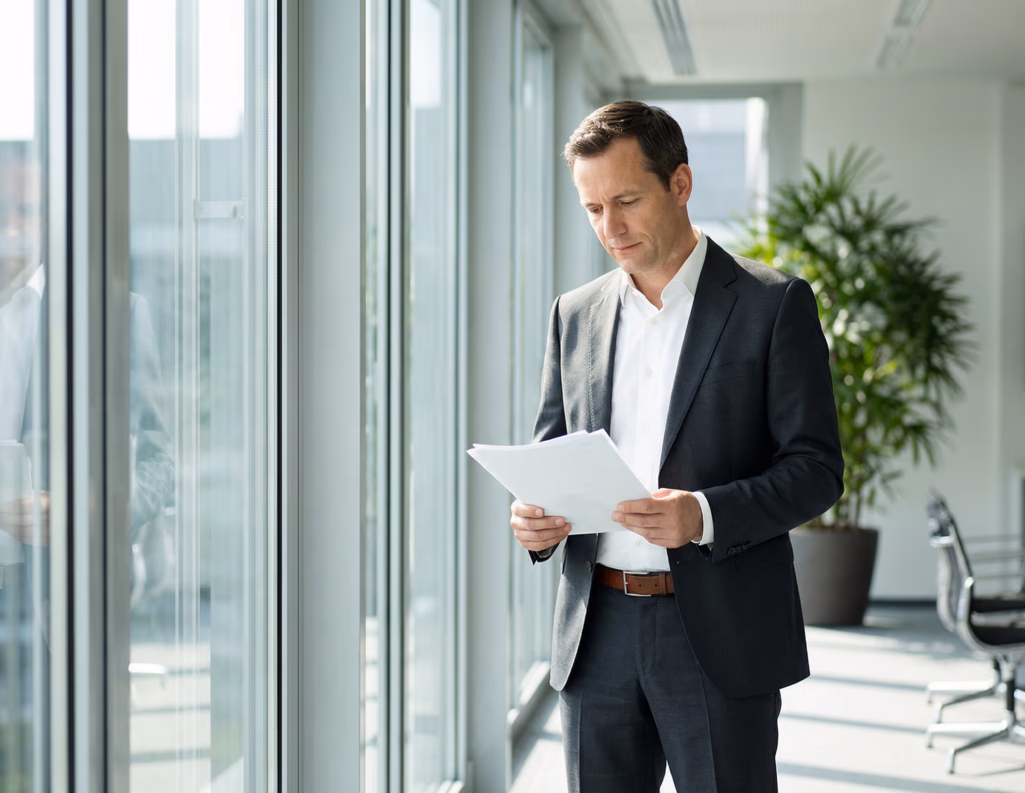 Man in a blue suit holding and reading papers near a window in an office setting.