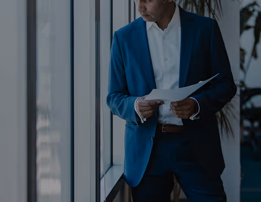 Man in a blue suit holding and reading papers near a window in an office setting.