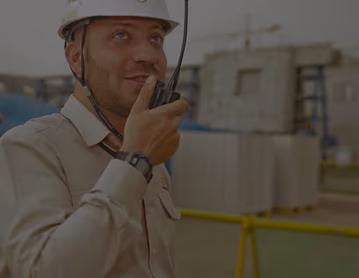Construction worker wearing a white helmet speaking into a walkie-talkie at a construction site.