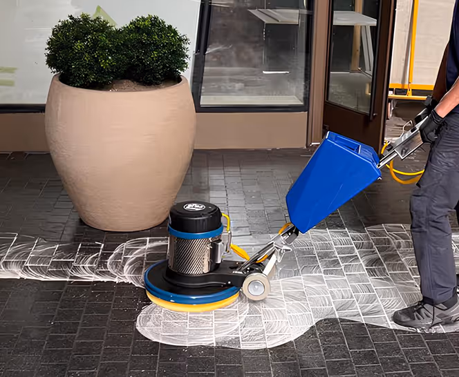 Person using a blue floor polishing machine on dark tiled flooring next to a large beige planter with green bushes.