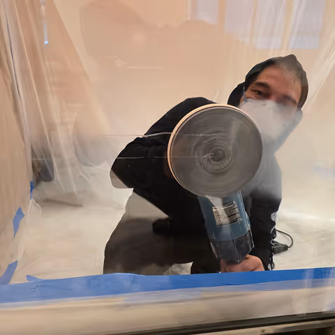 Person wearing mask and black hoodie polishing or grinding a glass surface indoors.