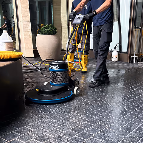 Two workers cleaning dark tiled floor using an electric floor scrubber machine.