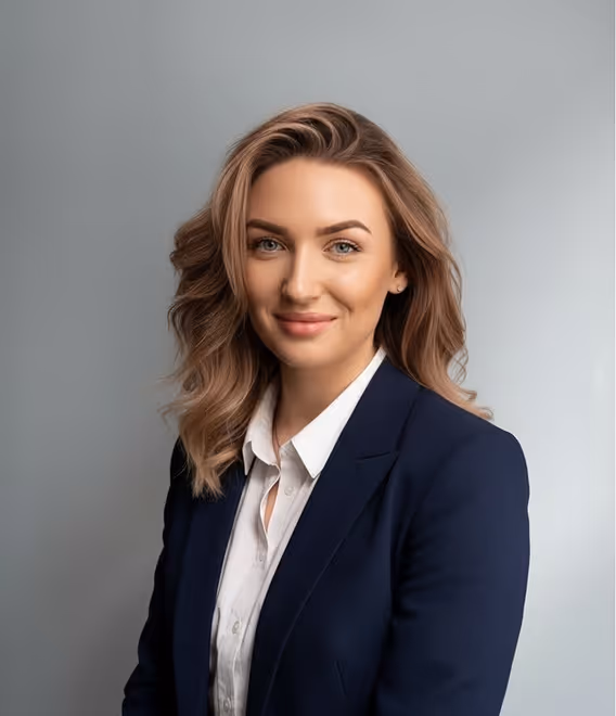 Smiling woman with wavy light brown hair wearing a navy blazer and white shirt against a gray background.
