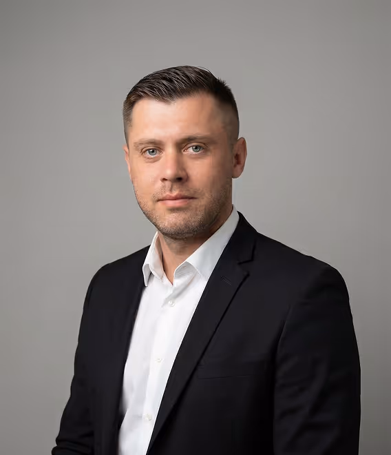 Man with short hair wearing a black blazer and white shirt against a gray background.