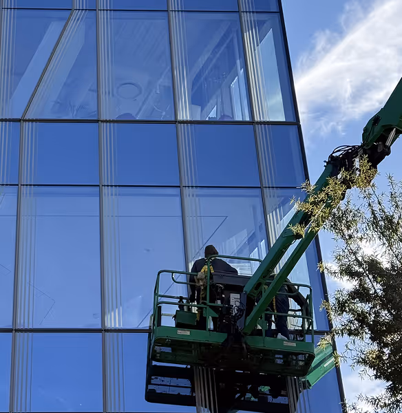 Person cleaning or inspecting glass windows on a tall building from a green aerial lift platform.