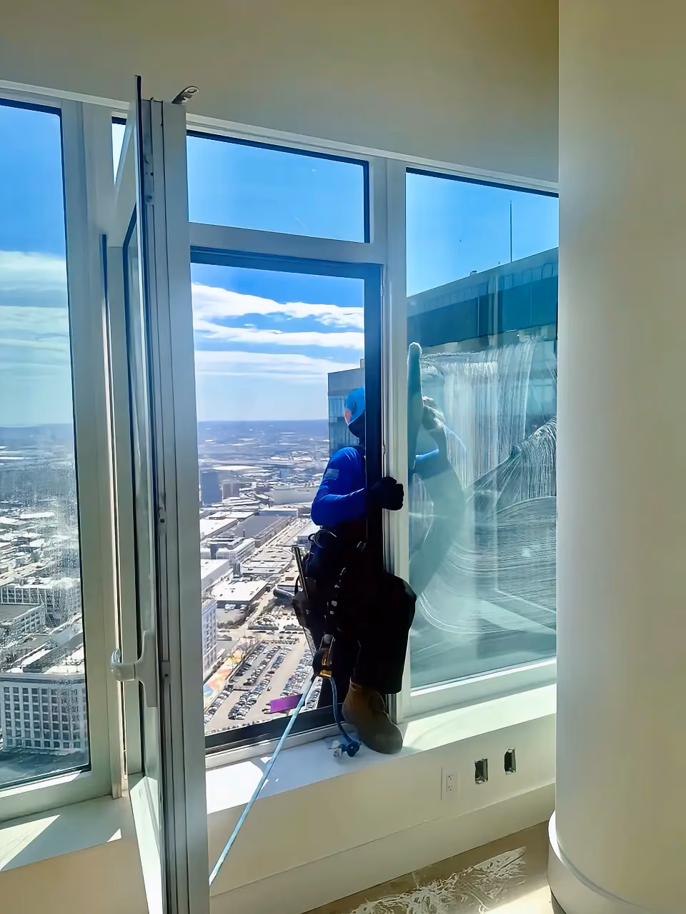 Worker in safety gear cleaning the outside of a high-rise building window with city view in the background.