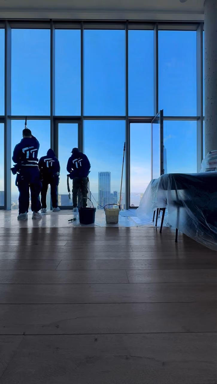 Three workers in uniforms with tools cleaning large floor-to-ceiling windows overlooking a city skyline.