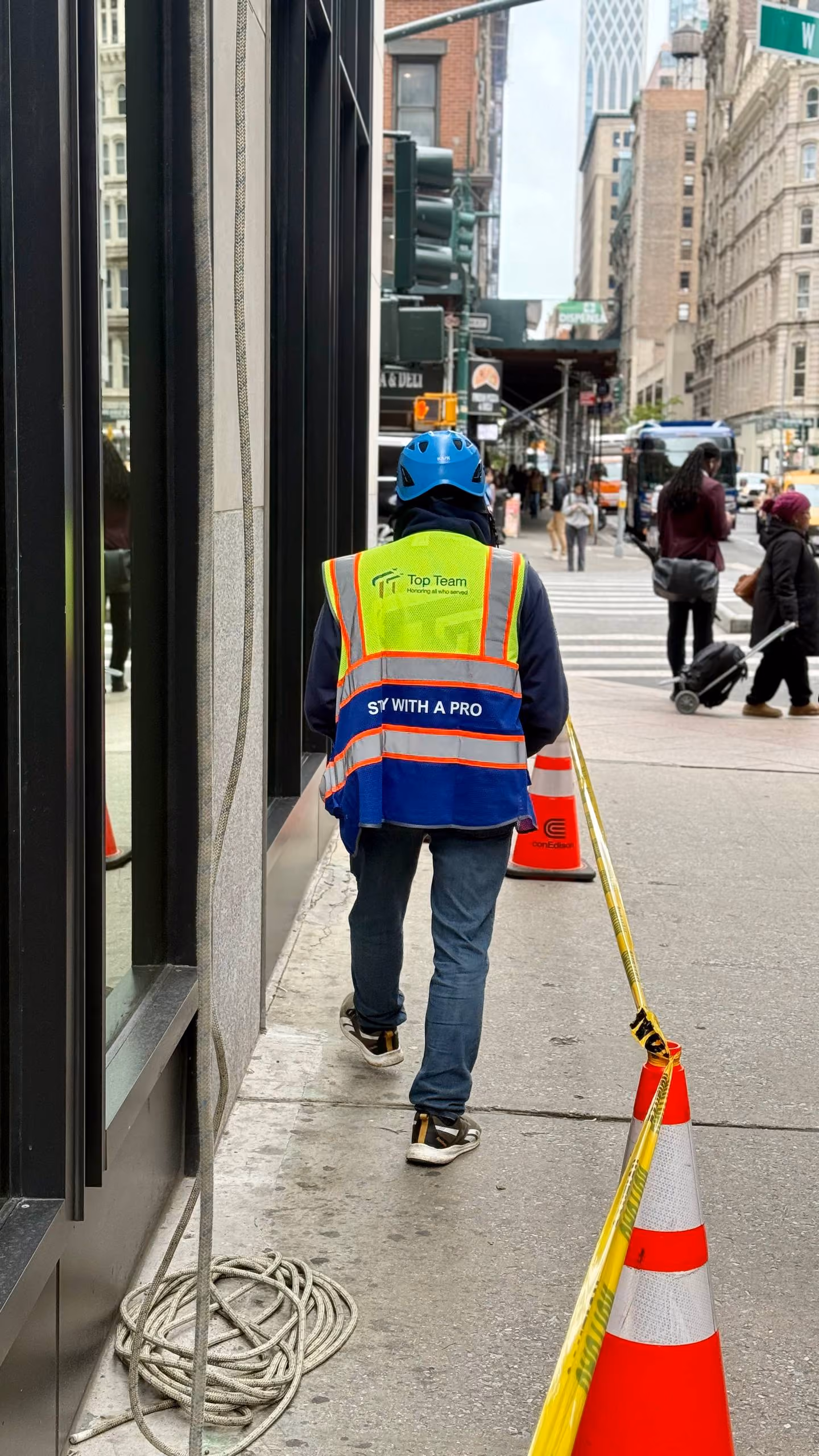 Worker wearing a Top Team safety vest and blue helmet walking on a city sidewalk near traffic cones and caution tape.
