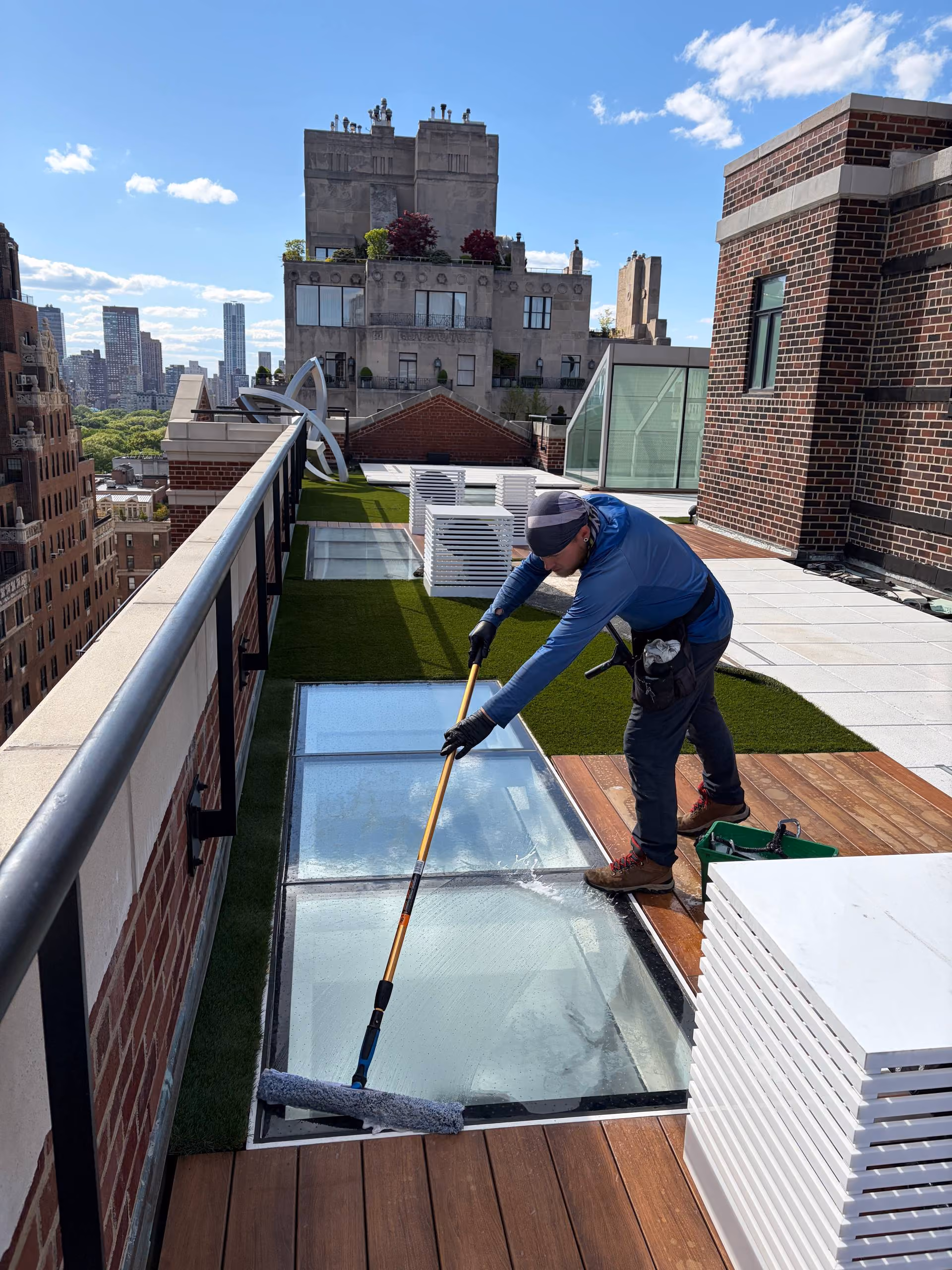 Worker cleaning large glass panels on a rooftop deck with wood and artificial grass under a sunny sky.