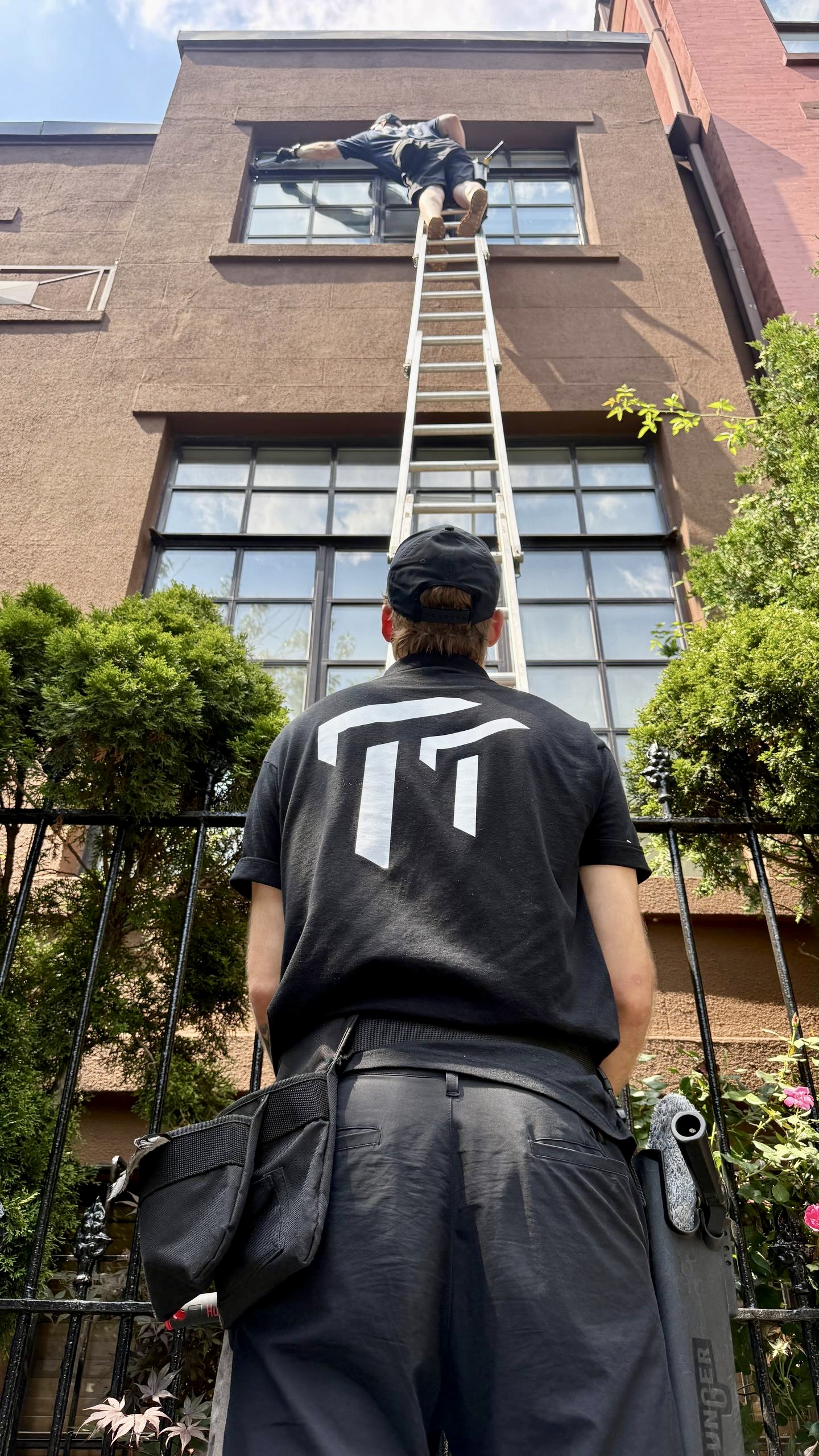 Worker in black watches colleague cleaning second-story window from tall ladder against brick building.