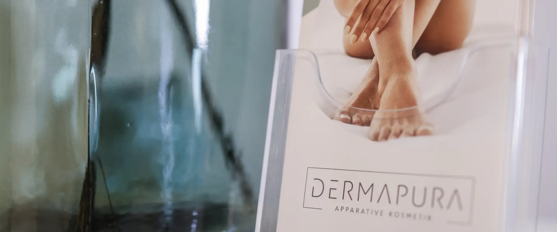Nahaufnahme von gepflegten Füßen und Händen einer sitzenden Person neben einem transparenten Behälter mit dem Schriftzug Dermapura Apparative Kosmetik.