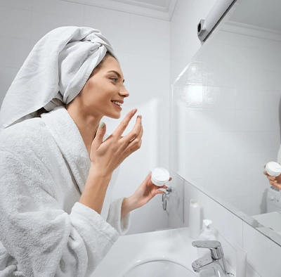 Frau im Bademantel mit Handtuch auf dem Kopf trägt im Bad Gesichtscreme auf ihr Gesicht auf.