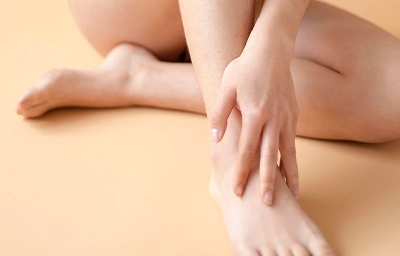 Frau sitzt mit verschränkten Beinen auf beigem Untergrund und hält eine Hand sanft am Knöchel.