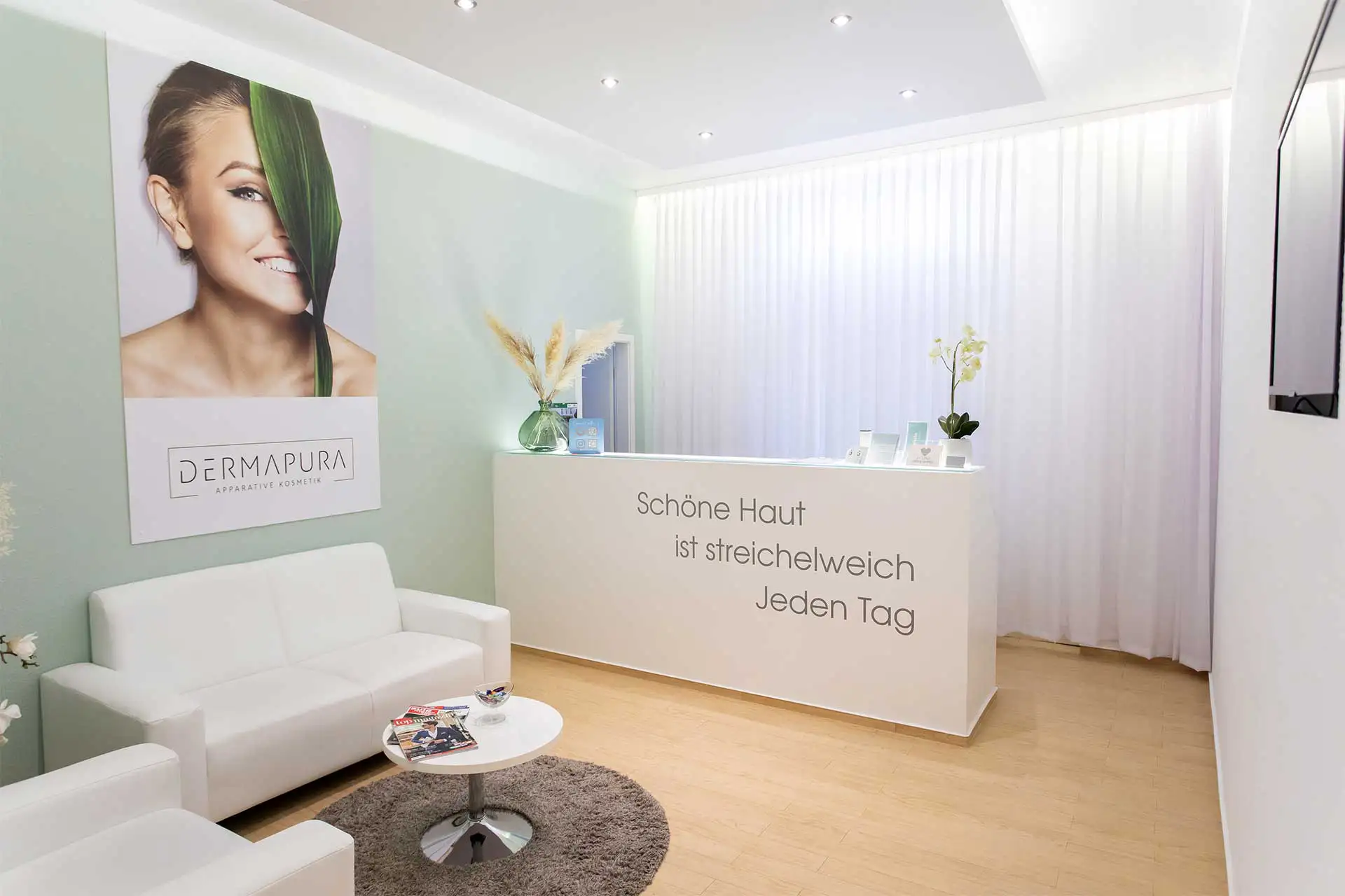 Helle Empfangs- und Wartebereich mit weißen Sofas, kleinem Tisch, Pflanzen und Theke mit Schriftzug 'Schöne Haut ist streichelweich Jeden Tag'.