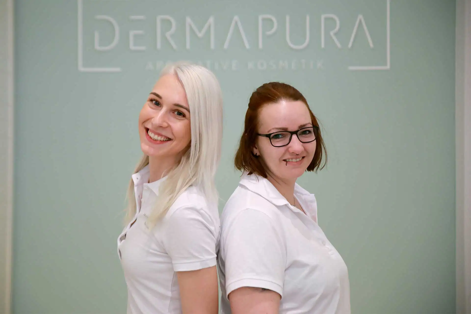 Zwei Frauen in weißen Poloshirts stehen Rücken an Rücken lächelnd vor einer Wand mit der Aufschrift Dermapura.