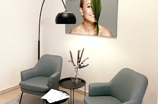 Gemütliche Sitzecke mit zwei grauen Sesseln, einem kleinen runden Tisch mit Vase und einer Stehlampe vor einer Wand mit einem Porträt einer lächelnden Frau, deren Gesicht teils von einem grünen Blatt verdeckt ist.