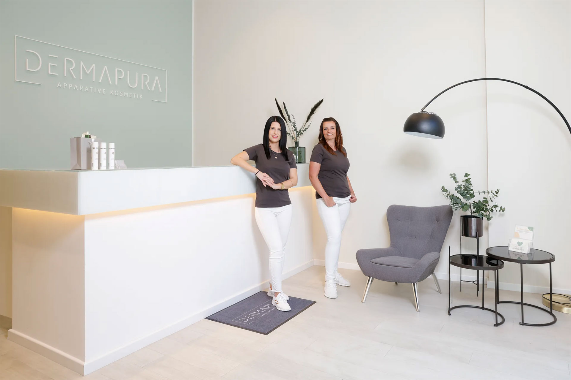 Zwei Frauen in weißer Hose und grauem T-Shirt lehnen an einem weißen Empfangstresen in einem modernen Kosmetikstudio.