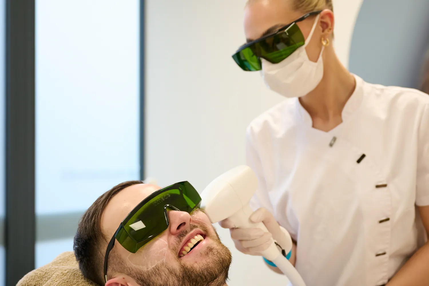 Mann erhält eine Laser-Haarentfernungsbehandlung im Gesicht durch eine Fachkraft mit Schutzbrille und Maske.
