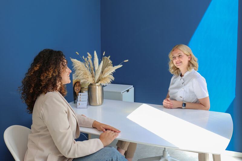 Zwei Frauen sitzen sich an einem weißen Tisch in einem Raum mit blauer Wand gegenüber und führen ein Gespräch.