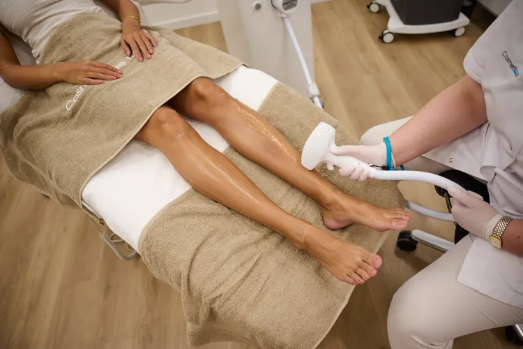 Frau liegt auf Behandlungsliege, während eine Kosmetikerin eine Laser-Haarentfernung an ihren glatten Beinen durchführt.