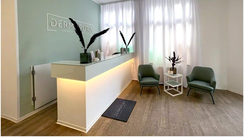 Moderner Empfangsbereich mit beleuchtetem weißen Tresen, zwei grünen Sesseln, kleinem Tisch und großen weißen Vorhängen.