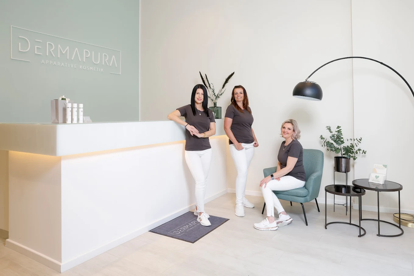 Drei Frauen in weißer Hose und grauem T-Shirt in einem modernen Kosmetikstudio mit dem Logo Dermapura an der Wand.