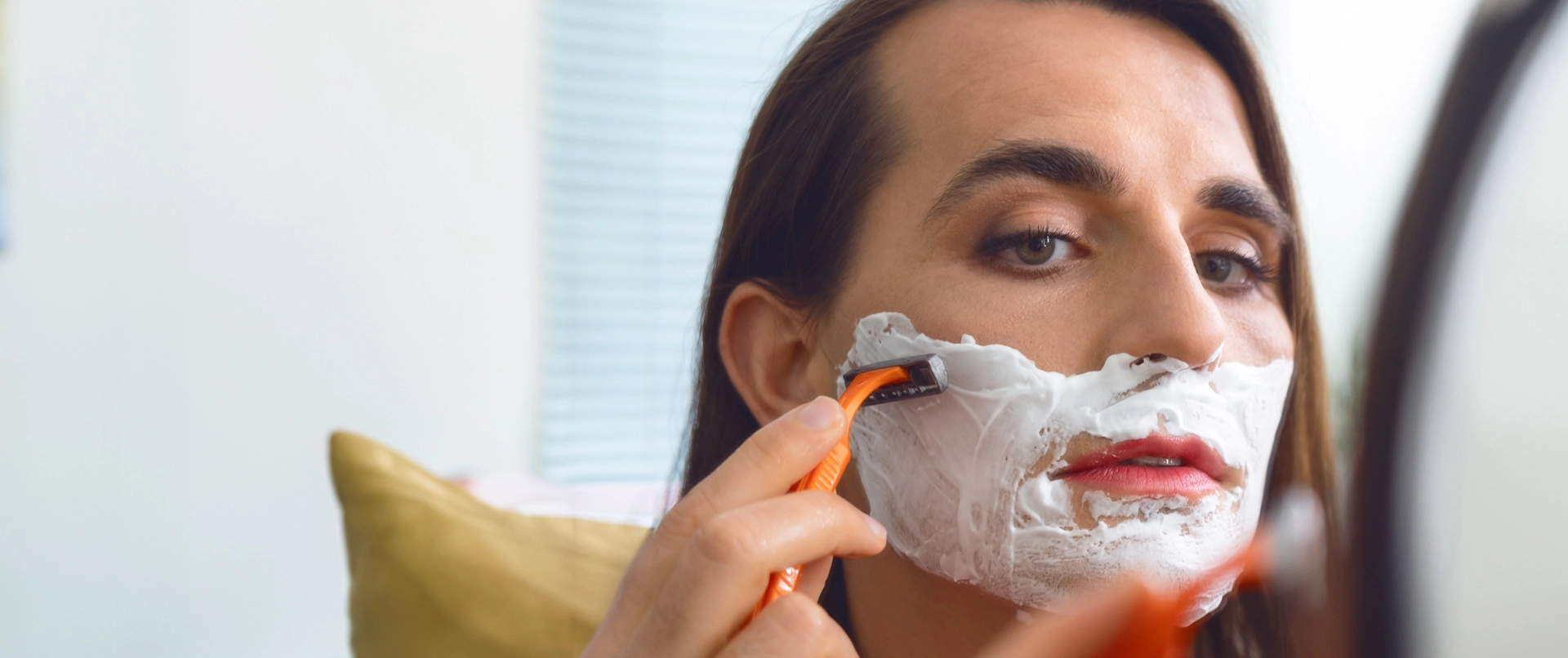 Person mit langen Haaren rasiert ihr Gesicht mit Rasiercreme und einem orangefarbenen Rasierer vor einem Spiegel.