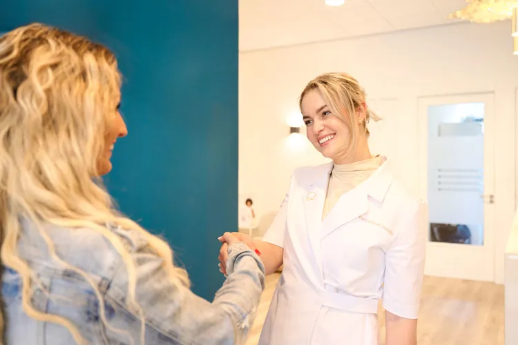 Eine Frau im weißen Kittel begrüßt lächelnd eine Kundin mit Handschlag in einer hellen Behandlungs- oder Empfangsumgebung.