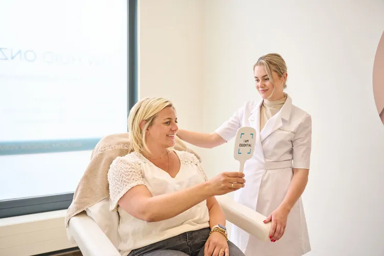 Eine Frau sitzt in einem Stuhl eines Kosmetikstudios und betrachtet lächelnd einen Handspiegel mit der Aufschrift 'I AM ESSENTIAL', während eine Kosmetikerin sie freundlich berührt.